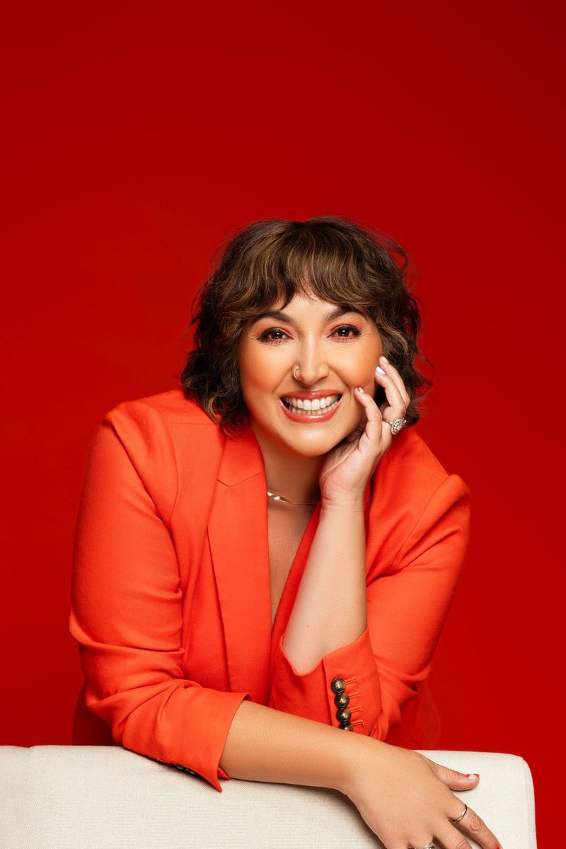Rachel Dee Minyard smiling at the camera, leaning on the back of a white chair in an orange blazer against a red backdrop