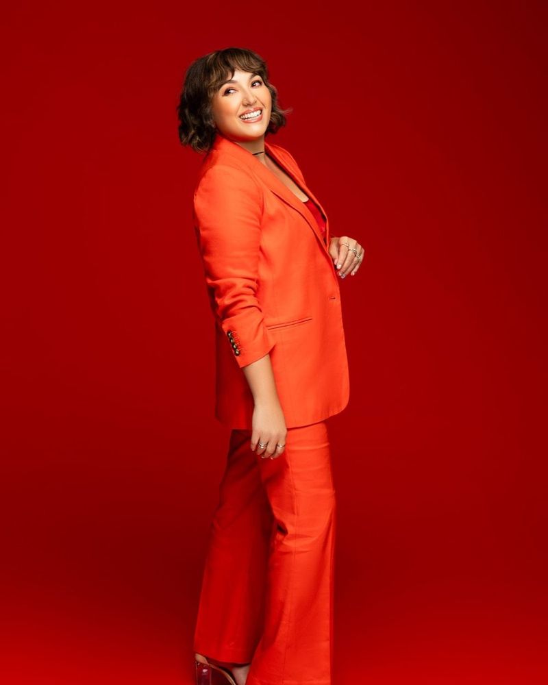 Rachel Dee Minyard standing in profile in an orange suit, smiling and looking back over her shoulder, against a deep red backdrop