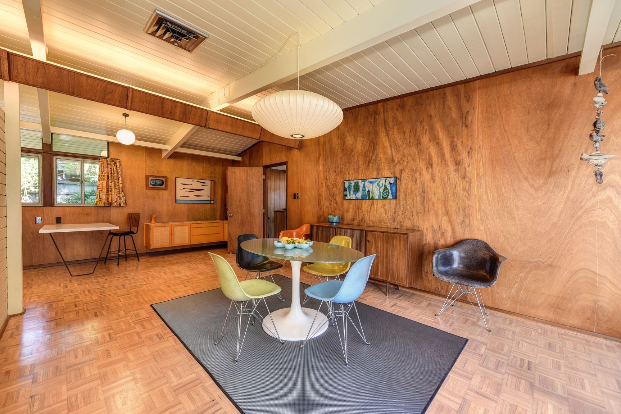 An open mid-century living and dining room with a beamed ceiling, mahogany paneling and molded-plastic chairs
