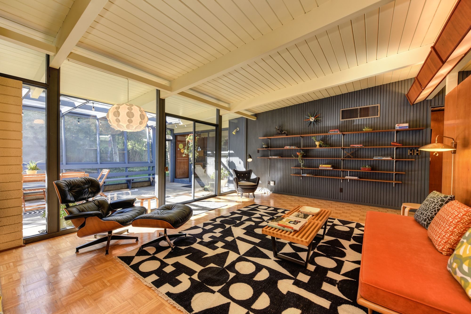 A mid-century modern living room with exposed beams, wood paneling, a wall of glass and a lounge chair