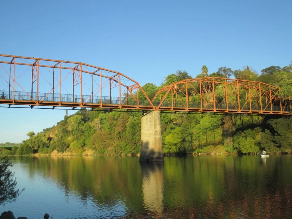 The orange steel Fair Oaks Bridge over the American River at golden hour, with a boat on the water